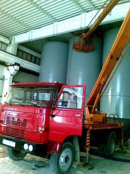 Czerwony samochód ciężarowy STAR z podnośnikiem koszowym na tle dużych, szarych zbiorników w hali przemysłowej; otwarte drzwi kabiny kierowcy.