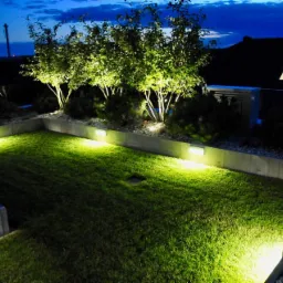 Oświetlony trawnik w ogrodzie nocą z niskimi, kwadratowymi lampami wzdłuż betonowego murku i drzewami podświetlonymi od dołu, widok z góry na posesję.