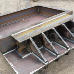 Spawana konstrukcja stalowa z profili zamkniętych i płaskowników, widoczne spoiny, surowy metal z rdzawym nalotem, na betonowym podłożu.