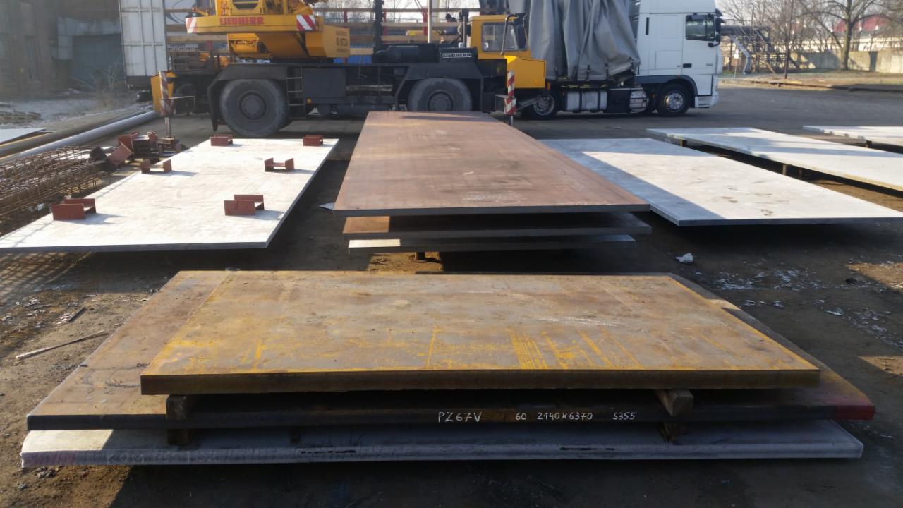 Stalowe płyty o różnych wykończeniach, z widocznym dźwigiem Liebherr i ciężarówką w tle, sugerujące przygotowanie do budowy konstrukcji.