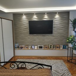 Usługi Remontowo-Budowlane Mateusz Pacuła - Nowoczesny salon z dekoracyjną ścianą 3D, podświetleniem LED, telewizorem, ramkami ze zdjęciami na półce i roślinami doniczkowymi na metalowym stojaku.