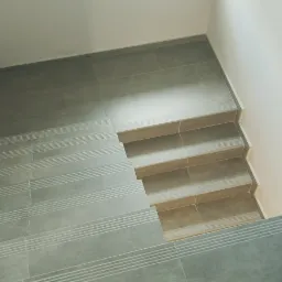 Widok z góry na wyłożone szarymi płytkami schody z delikatnymi, poziomymi rowkami antypoślizgowymi, prowadzące w dół, z białymi ścianami po bokach.