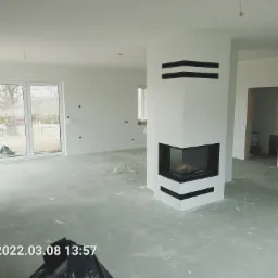 Wnętrze w trakcie remontu: widoczne ślady gładzi na podłodze, nowoczesny biały kominek z czarnymi akcentami, duże okna z widokiem na zewnątrz, puszka farby w rogu pomieszczenia.