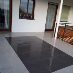 Usługi Remontowo-Budowlane Mateusz Pacuła - Wyłożony płytkami taras z kontrastującym wzorem ciemnych i jasnych płytek, drewniana balustrada, widok na budynek z oknami.