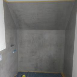 Usługi Remontowo-Budowlane Mateusz Pacuła - Szara, surowa faktura ścian w łazience, zabezpieczone żółtą taśmą malarską przy suficie i podłodze, widoczna wnęka w ścianie oraz wystający fragment instalacji wodnej.