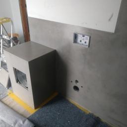 Usługi Remontowo-Budowlane Mateusz Pacuła - Szara ściana z gładzią dekoracyjną, widoczne otwory pod instalacje i puszka elektryczna, obudowa pod geberit z otworem rewizyjnym, zabezpieczona żółtą taśmą malarską.