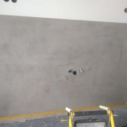 Usługi Remontowo-Budowlane Mateusz Pacuła - Ściana pokryta szarą, nieregularną fakturą gładzi, widoczne otwory na gniazdka elektryczne i rury, zabezpieczona podłoga żółtą taśmą malarską, lampa robocza i odkurzacz w tle.