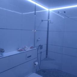 Usługi Remontowo-Budowlane Mateusz Pacuła - Nowoczesna łazienka z białymi płytkami, podświetleniem LED i szklaną kabiną prysznicową typu walk-in, z widocznymi akcesoriami łazienkowymi na półce.