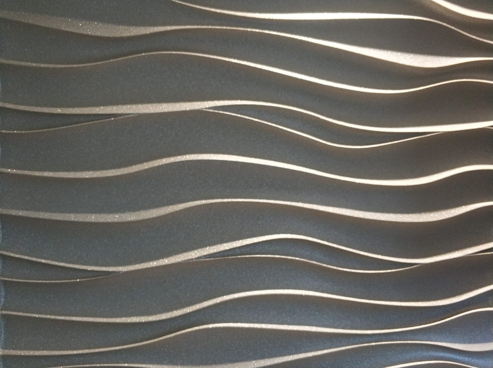 Tekstura ściany z falistymi, trójwymiarowymi panelami w odcieniach szarości z delikatnym połyskiem, tworzącymi efekt głębi i ruchu.