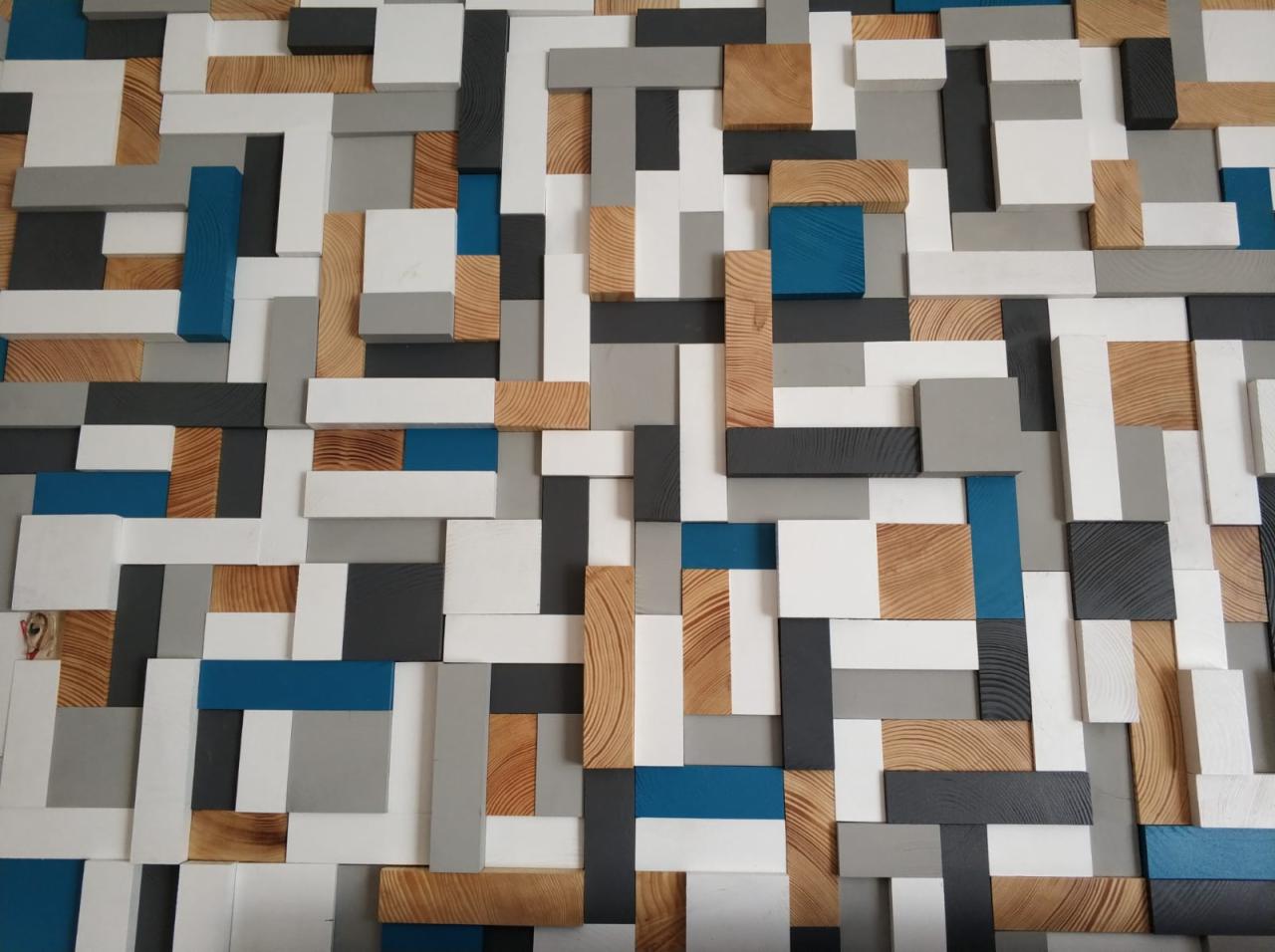 Dekoracyjna ściana z drewnianych i malowanych na biało, szaro i niebiesko klocków o różnych rozmiarach, tworząca trójwymiarową mozaikę.