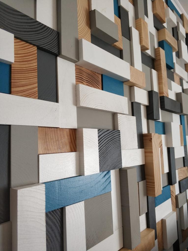 Dekoracyjna ściana z drewnianych, trójwymiarowych elementów w odcieniach bieli, szarości, naturalnego drewna i błękitu, tworząca geometryczny wzór.