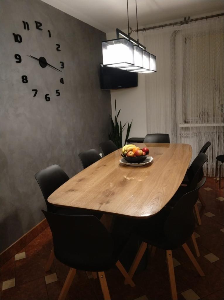 Jadalnia z drewnianym stołem, czarnymi krzesłami, owocami w misie, zegarem ściennym i nowoczesnym oświetleniem wiszącym.