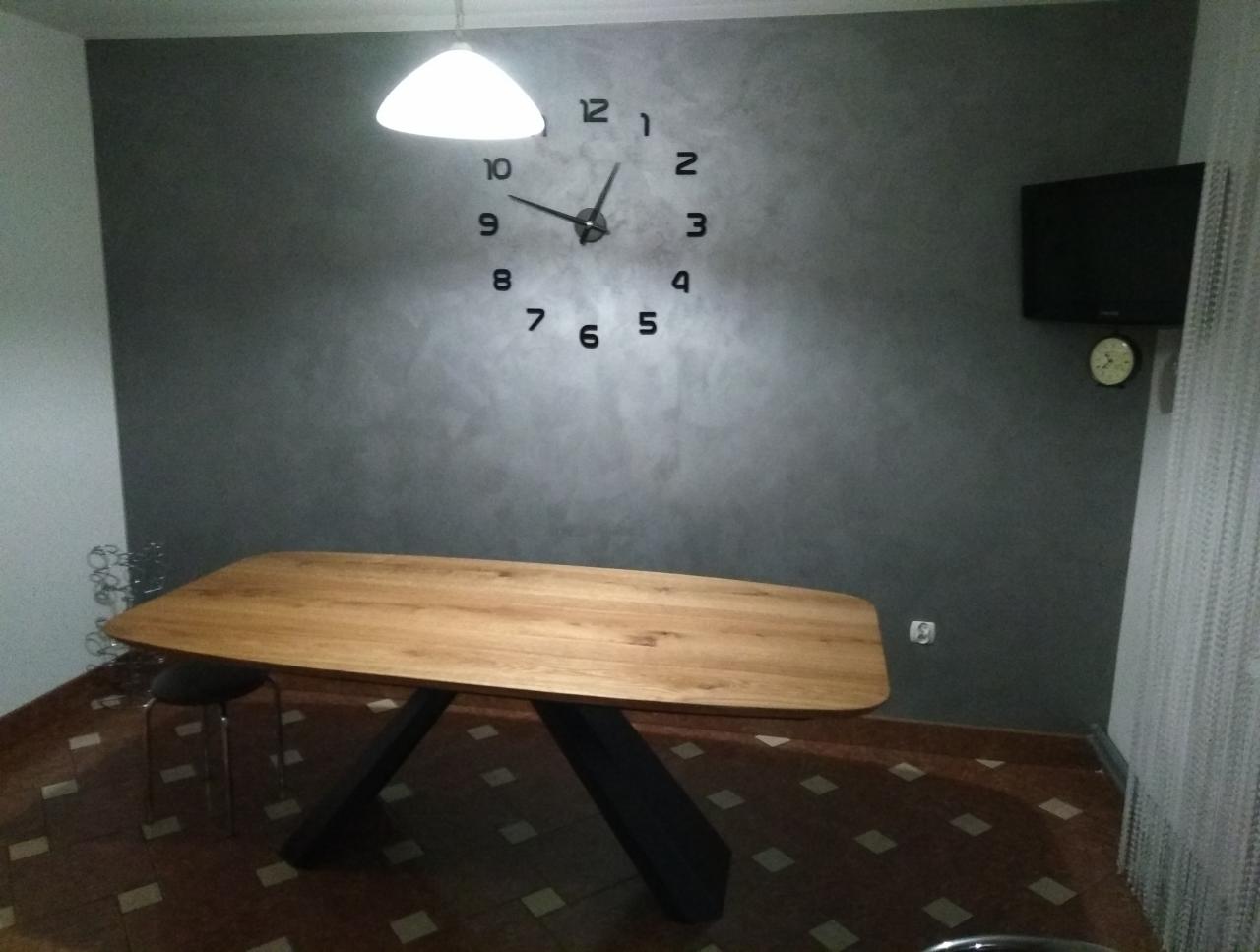 Szara ściana z dekoracyjną gładzią w salonie, ozdobiona dużym, naklejanym zegarem i telewizorem, oświetlona lampą sufitową, z drewnianym stołem na pierwszym planie.