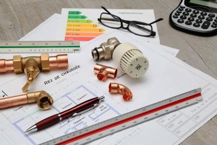 Plan instalacji grzewczej z miedzianymi złączkami, zaworem termostatycznym, kalkulatorem, okularami i długopisem na drewnianym biurku, widok z góry.