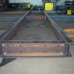 Fragment konstrukcji stalowej z widocznymi otworami i śrubami, spawarka i wózek widłowy w tle.