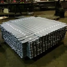 Stos srebrnych, metalowych elementów z zaokrąglonymi końcami, ułożonych warstwami w hali produkcyjnej, z widocznymi śladami obróbki i numerami seryjnymi na wierzchu.