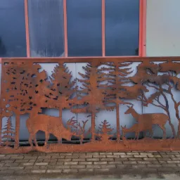 Dekoracyjny, rdzawy panel metalowy z wyciętym motywem leśnym: jelenie i drzewa, oparty o szarą ścianę budynku z czerwonymi ramami okiennymi, leżący na chodniku z kostki.