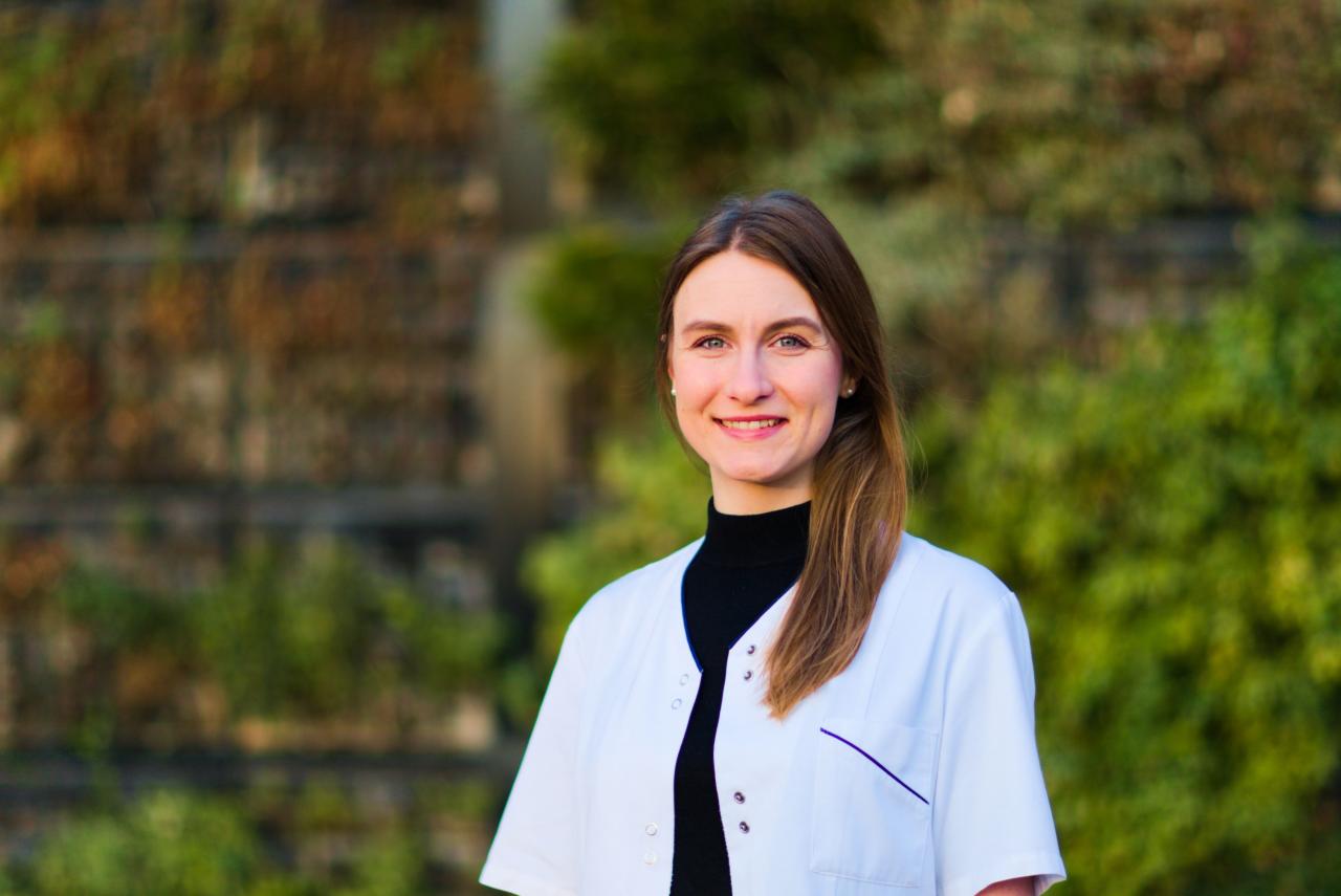 Uśmiechnięta dietetyczka w białym uniformie na tle zielonej ściany z roślin.