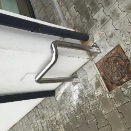 Metalowa poręcz zamontowana na elewacji budynku, widoczna z dołu, z fragmentem rynny i brukowanym chodnikiem z pokrywą studzienki kanalizacyjnej