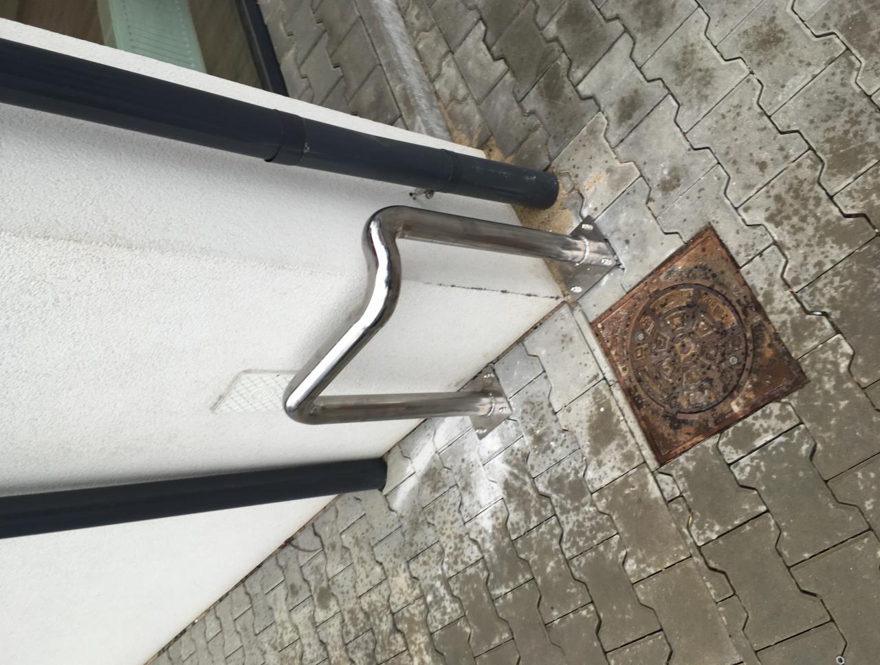 Metalowa poręcz zamontowana na elewacji budynku, widoczna z dołu, z fragmentem rynny i brukowanym chodnikiem z pokrywą studzienki kanalizacyjnej
