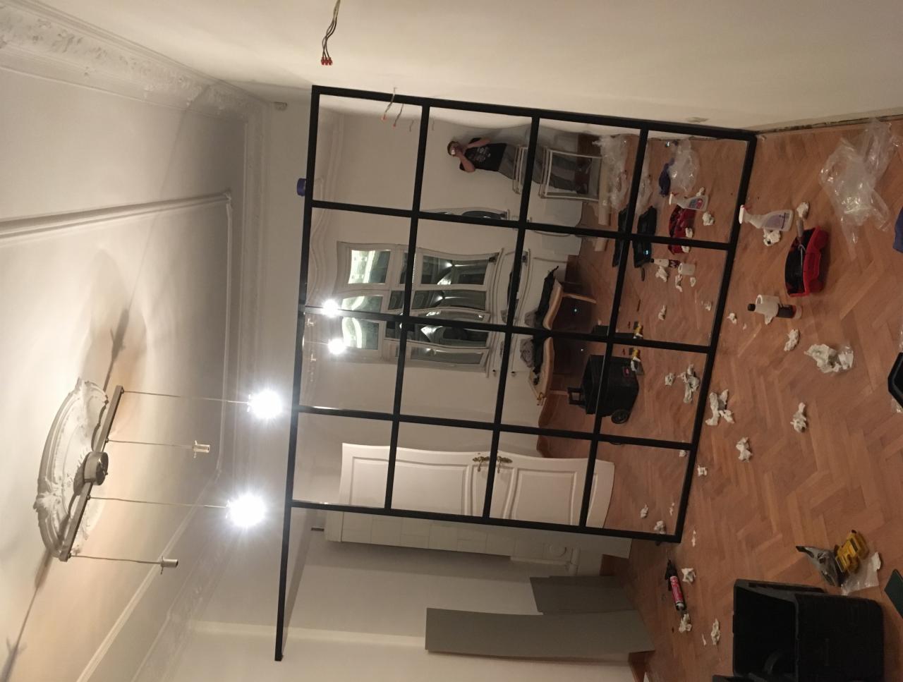 Montaż metalowej ścianki działowej z czarnymi profilami w jasnym pomieszczeniu; widoczne narzędzia i resztki materiałów na parkiecie.