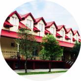 Elewacja budynku z czerwonym dachem i oknami mansardowymi, widoczna z poziomu trawnika, z zielenią w tle.