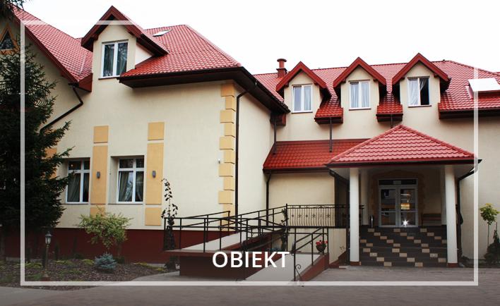 Elewacja budynku z czerwonym dachem i wejściem z rampą dla niepełnosprawnych, otoczona zielenią.