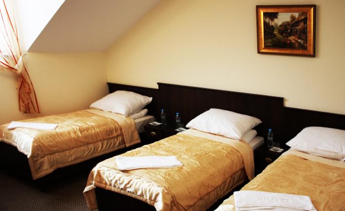 Pokój trzyosobowy z trzema pojedynczymi łóżkami z beżowymi narzutami, białymi poduszkami i ręcznikami, z ciemnobrązowym wezgłowiem, butelkami wody mineralnej na szafkach nocnych i obrazem w złotej...