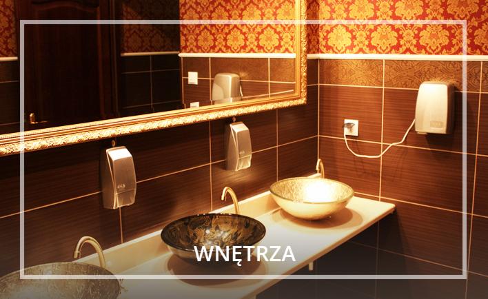 Elegancka łazienka z dwoma szklanymi umywalkami w odcieniach złota i brązu, ciemne płytki, ozdobne lustro w złotej ramie i wzorzysta tapeta.