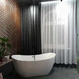 Nowoczesna łazienka z wolnostojącą białą wanną na tle okna z białymi i szarymi zasłonami, ściana pokryta dekoracyjnymi, trójwymiarowymi panelami w odcieniach brązu, ciemna podłoga z marmurowym...