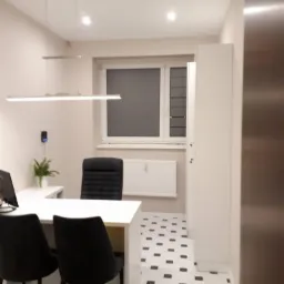 Nowoczesne biuro z białymi ścianami, czarnymi krzesłami i biało-czarną podłogą w karo, oświetlone podłużną lampą LED, z widocznym biurkiem i rośliną doniczkową.