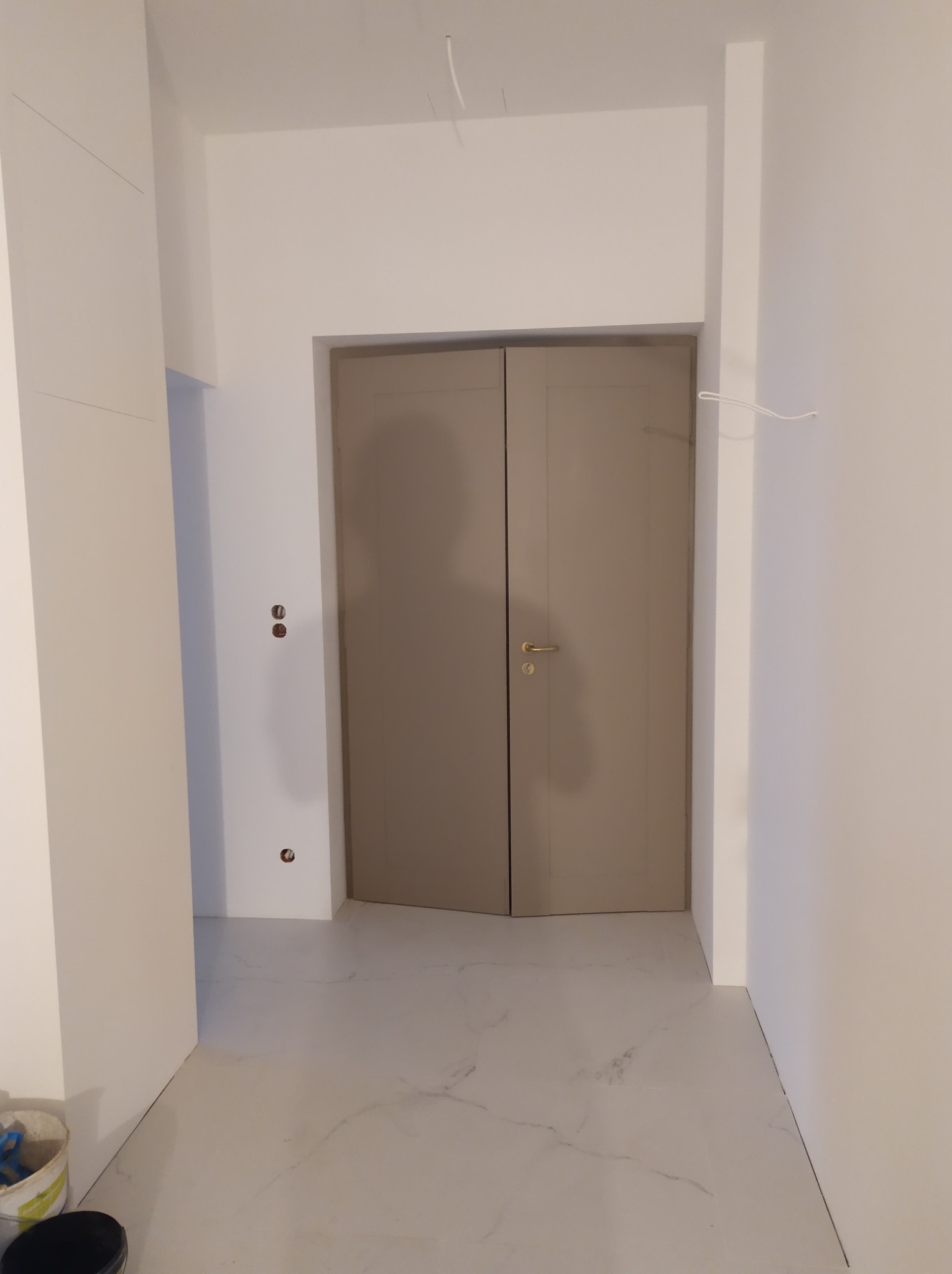 Nowe, dwuskrzydłowe drzwi wewnętrzne w odcieniach beżu z złotą klamką, zamontowane w odnowionym, jasnym pomieszczeniu z marmurową podłogą i świeżo pomalowanymi ścianami, widoczne niedokończone...