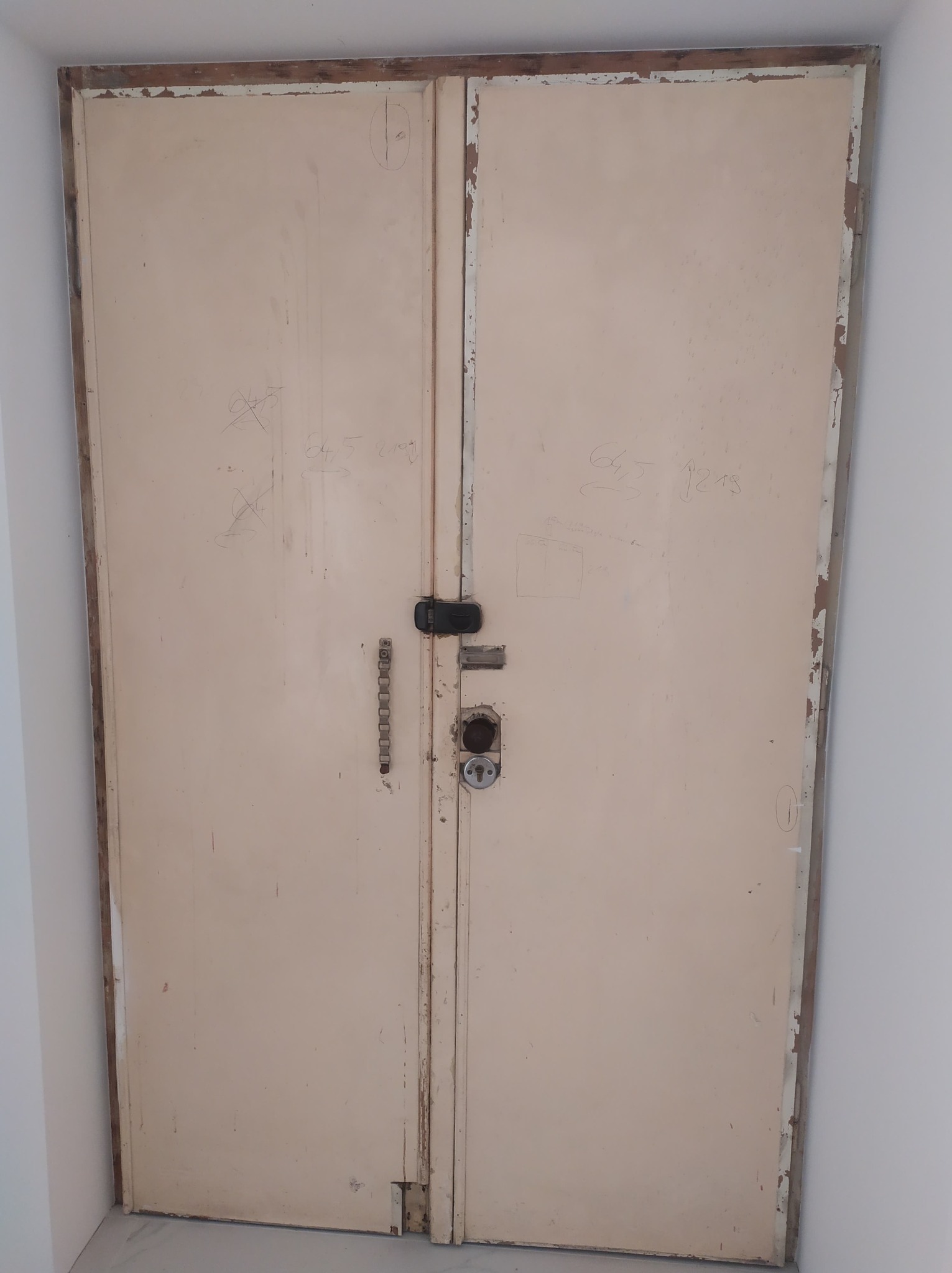 Stare, dwuskrzydłowe drzwi metalowe w kolorze beżowym, z widocznymi śladami użytkowania, rysami i napisami, z zamkiem i długim uchwytem, osadzone w białej ścianie.
