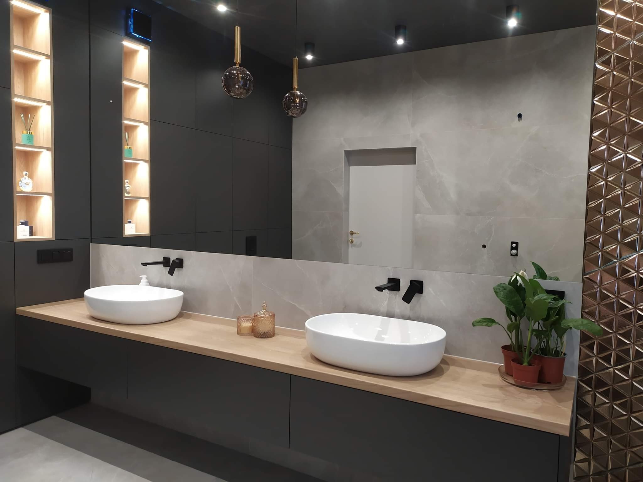 Nowoczesna łazienka z dwoma owalnymi umywalkami nablatowymi, czarną armaturą, szarą glazurą imitującą marmur i dekoracyjną ścianą z trójkątnych płytek w kolorze miedzianym, oświetlona wiszącymi...
