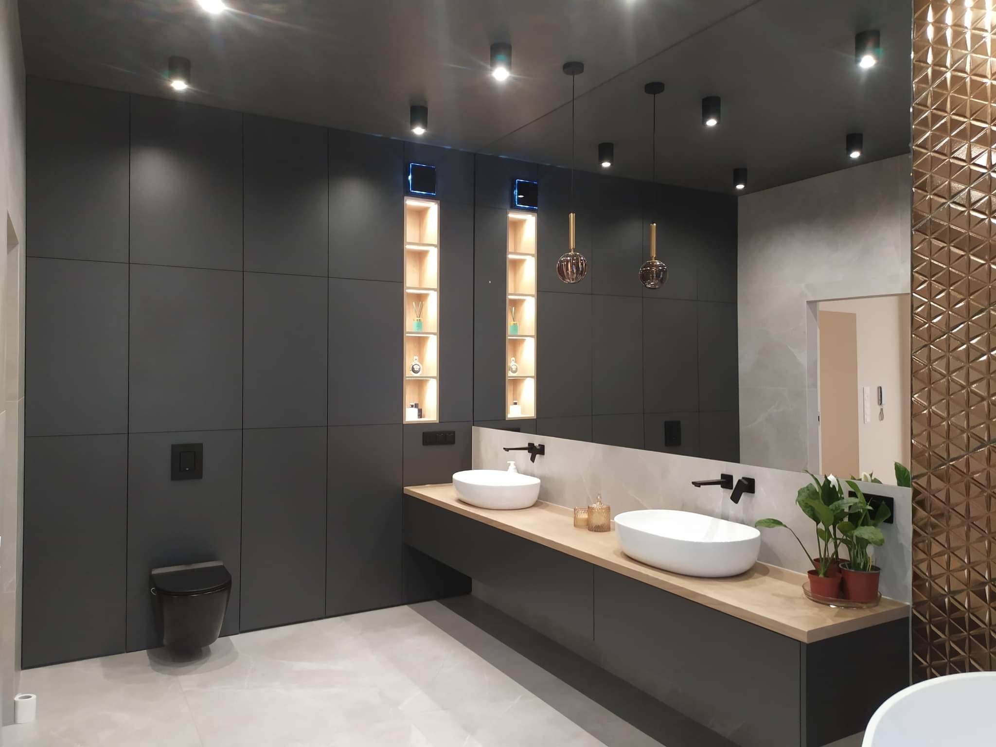 Nowoczesna łazienka z ciemnoszarymi panelami ściennymi, dwiema białymi umywalkami nablatowymi na drewnianym blacie, podwieszaną toaletą, dekoracyjnymi złotymi lamelami i oświetleniem punktowym