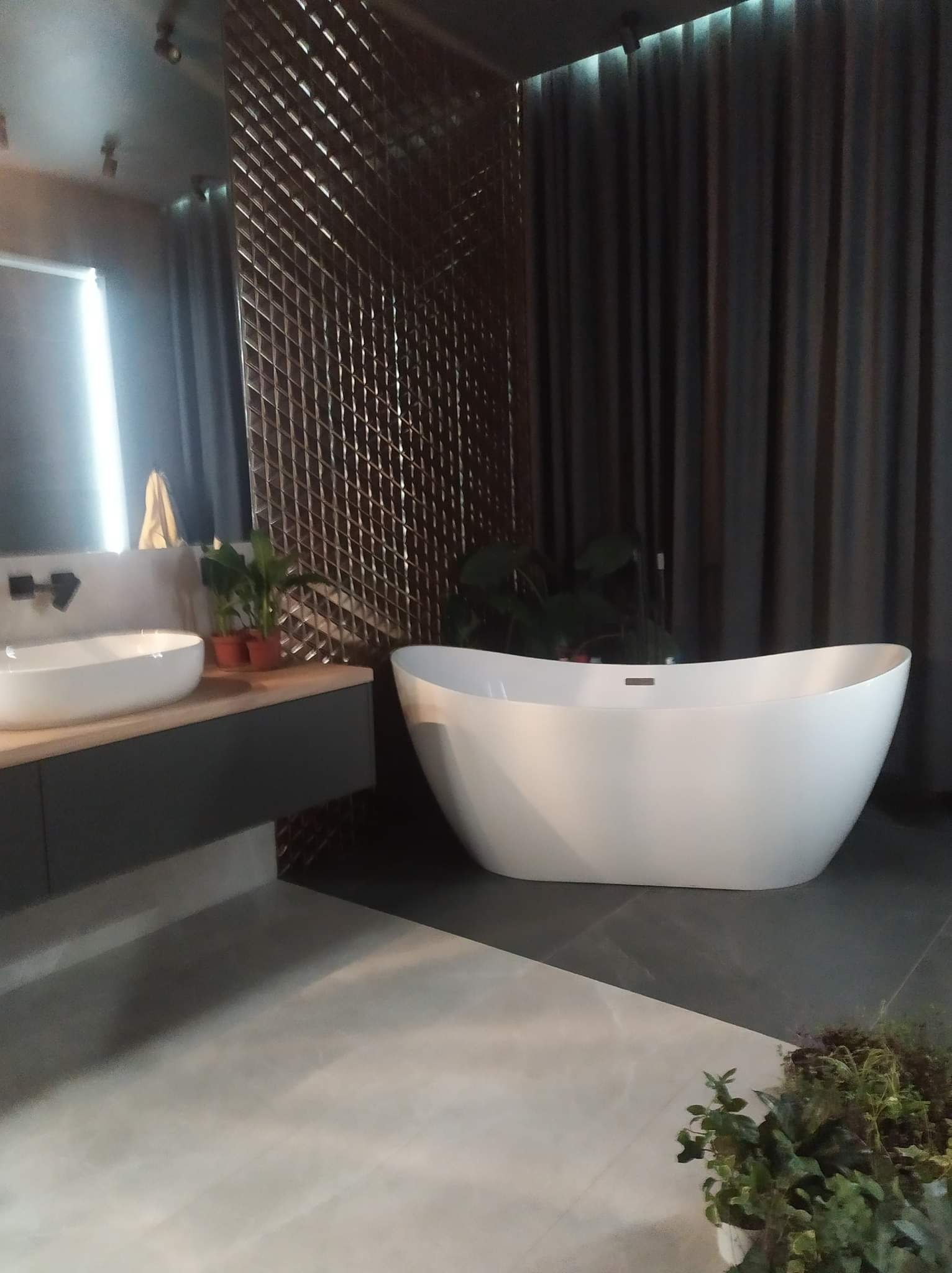 Nowoczesna łazienka z wolnostojącą białą wanną, umywalką nablatową na drewnianym blacie, szarą mozaiką na ścianie i ciemnymi zasłonami w tle.