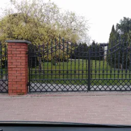Czarna, dwuskrzydłowa brama z kutego żelaza, osadzona na ceglanych słupkach, z widocznym trawnikiem i drzewami w tle.