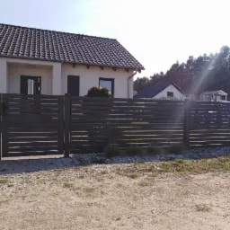 Nowoczesne, poziome przęsła ogrodzeniowe w kolorze antracytowym, z widocznym numerem domu '2' i skrzynką pocztową; tło stanowi dom jednorodzinny i las.