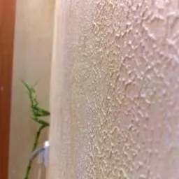 Beżowa ściana wewnętrzna z dekoracyjnym tynkiem o nieregularnej, chropowatej fakturze, oświetlona od góry, z fragmentem rośliny w lewym dolnym rogu.