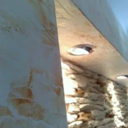 Dekoracyjna ściana z efektem tynku strukturalnego w ciepłych odcieniach beżu i brązu, połączona z fragmentem ściany z kamienia naturalnego, oświetlona punktowymi lampami sufitowymi.