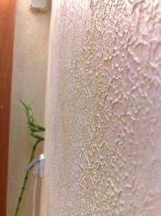 Beżowa ściana wewnętrzna z dekoracyjnym tynkiem o nieregularnej, chropowatej fakturze, oświetlona od góry, z fragmentem rośliny w lewym dolnym rogu.