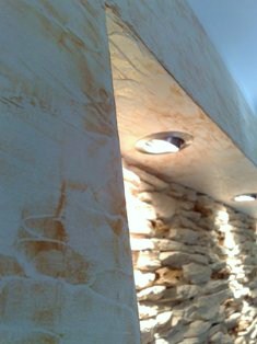 Dekoracyjna ściana z efektem tynku strukturalnego w ciepłych odcieniach beżu i brązu, połączona z fragmentem ściany z kamienia naturalnego, oświetlona punktowymi lampami sufitowymi.