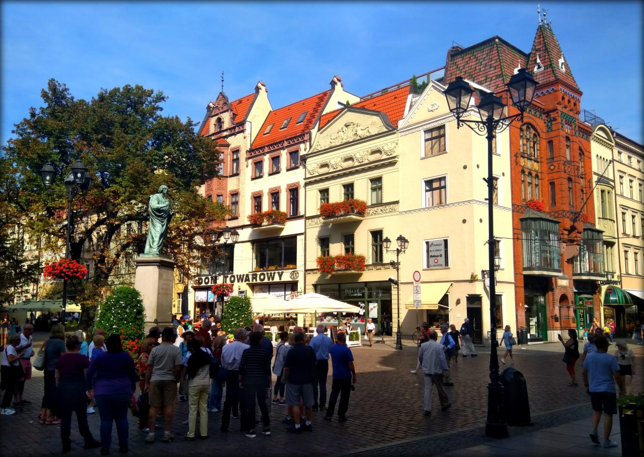 Rynek miejski z zabytkową architekturą, pomnikiem i zgromadzeniem ludzi w słoneczny dzień.
