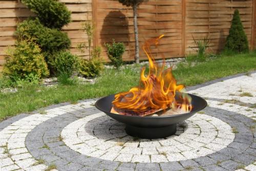 Ognisko w czarnej metalowej misie, otoczone okręgami z kostki brukowej na tle drewnianego ogrodzenia i zielonego trawnika.