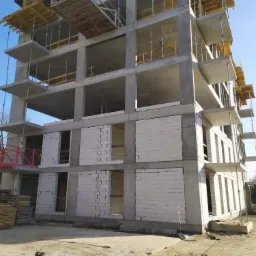 Wielopiętrowy budynek w trakcie budowy, widoczne betonowe szkielety i częściowo wymurowane ściany z białych bloczków, szalunki na górnych kondygnacjach, teren budowy z piaskiem i drewnianymi...