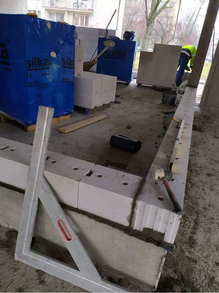 Wznoszenie ściany z białych bloczków Silka na zaprawie, widoczny poziomica, kątownik i palety z zapakowanymi bloczkami w tle.