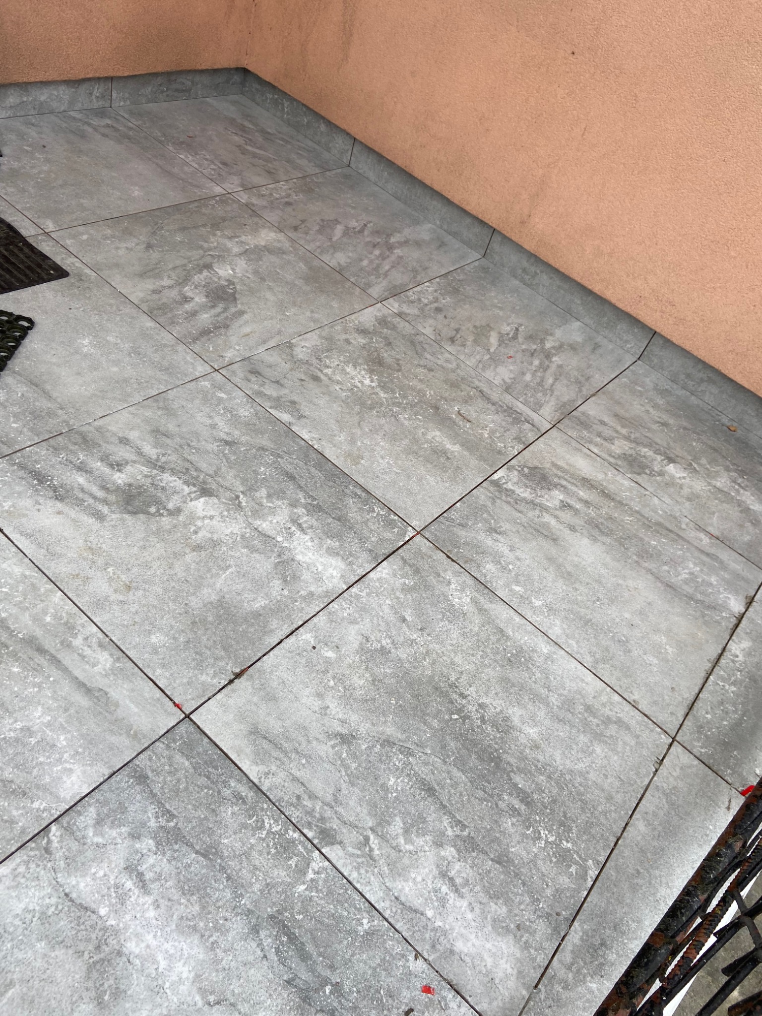 Szare, kwadratowe płytki podłogowe z delikatnym wzorem ułożone na zewnątrz, przylegające do ściany o nierównej fakturze i brzoskwiniowym odcieniu, z fragmentem czarnej, metalowej barierki w rogu.