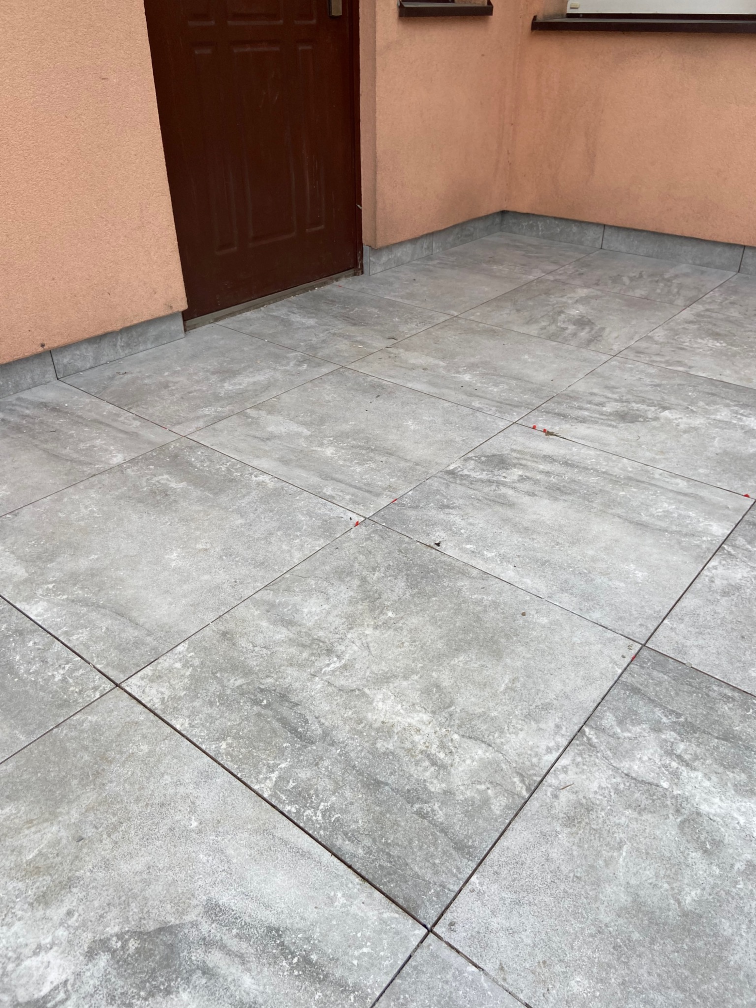 Szare płytki gresowe ułożone na zewnątrz budynku, widoczne czerwone kliny dystansowe w spoinach, brązowe drzwi wejściowe i fragment elewacji w tle.