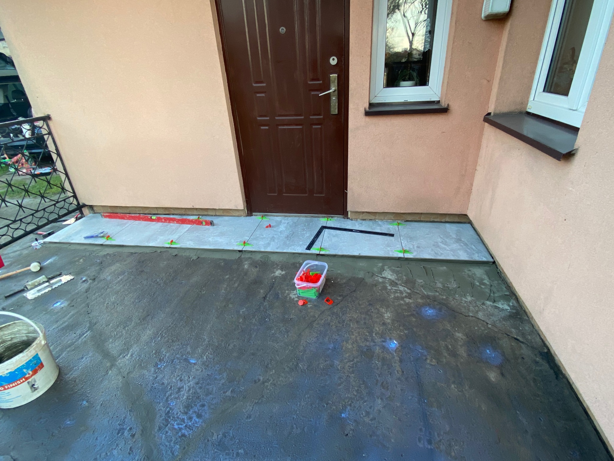 Kładzenie płytek na zewnątrz budynku przed drzwiami wejściowymi, widoczne poziomica, szpachelka, wiadro z klejem i pojemnik z klipsami poziomującymi.