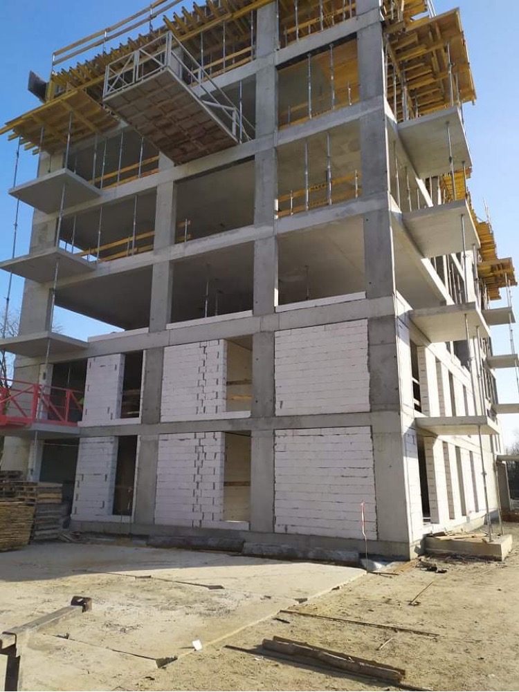 Wielopiętrowy budynek w trakcie budowy, widoczne betonowe szkielety i częściowo wymurowane ściany z białych bloczków, szalunki na górnych kondygnacjach, teren budowy z piaskiem i drewnianymi...